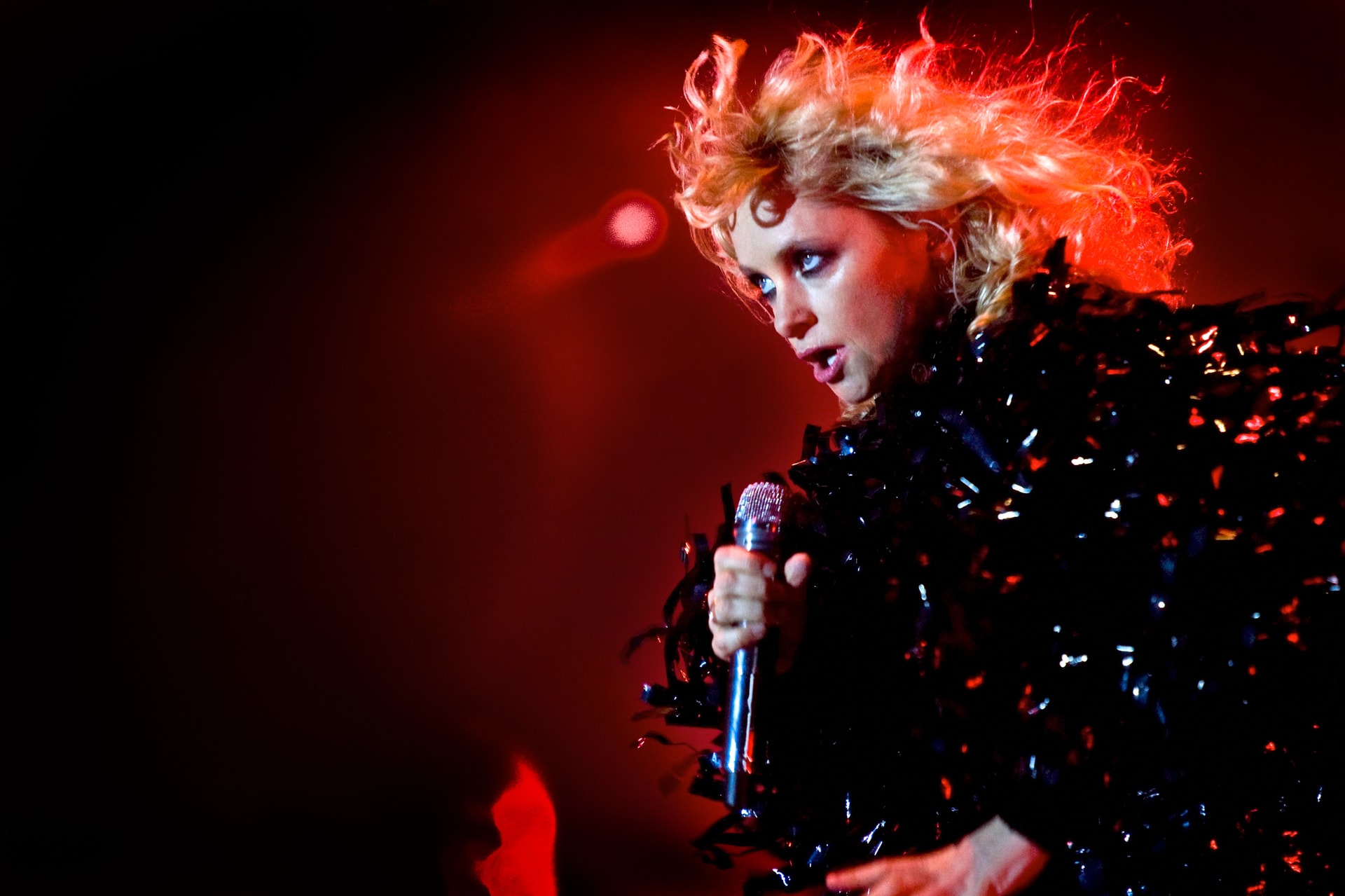 Alison Goldfrapp performing live with a microphone under dramatic red stage lighting