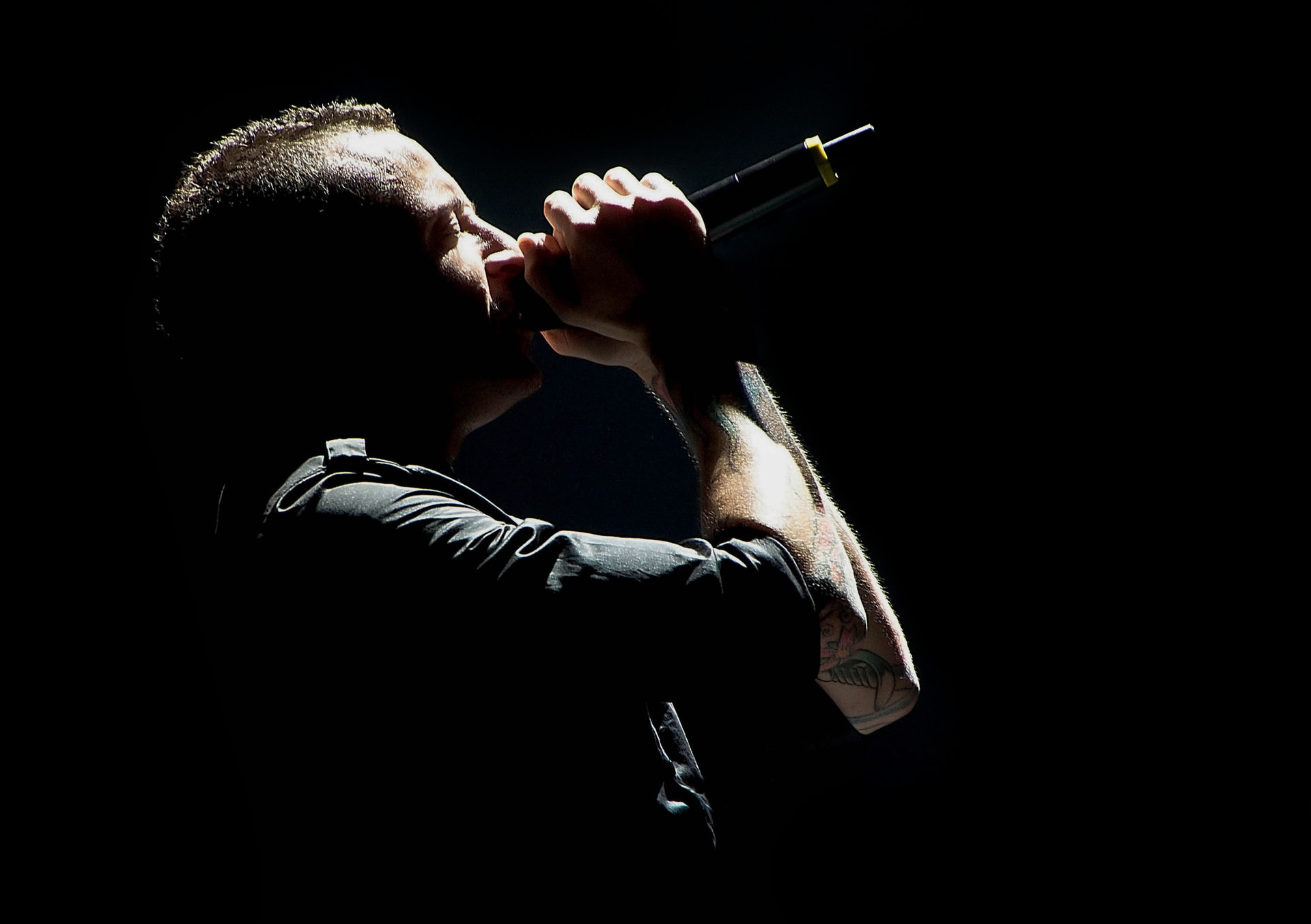 Chester Bennington of Linkin Park singing into a microphone, head bowed, lit dramatically against a black background