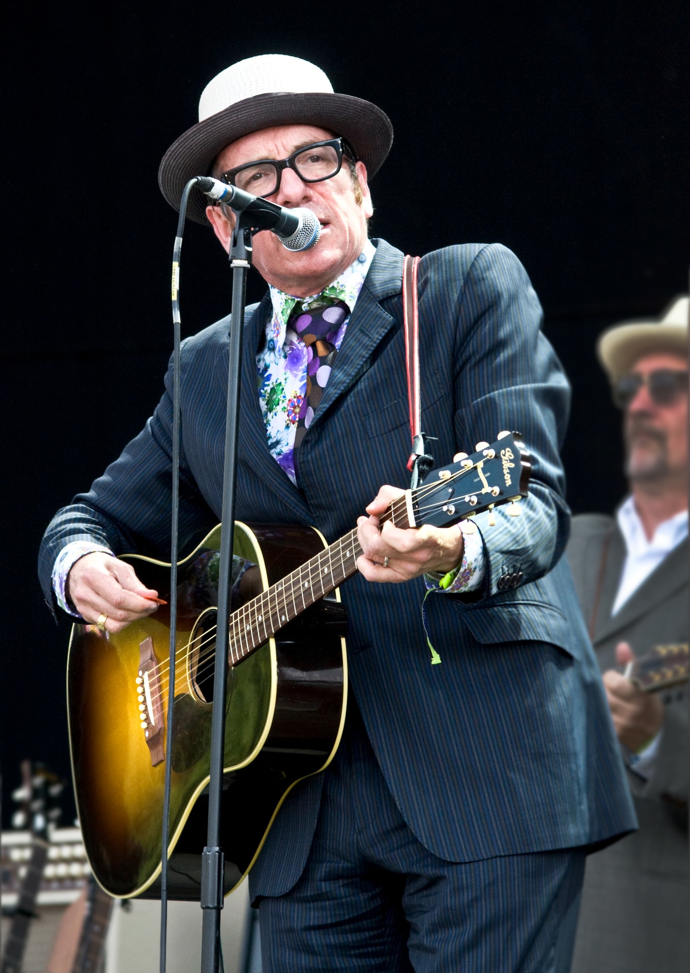 Elvis Costello performing live with an acoustic guitar and microphone