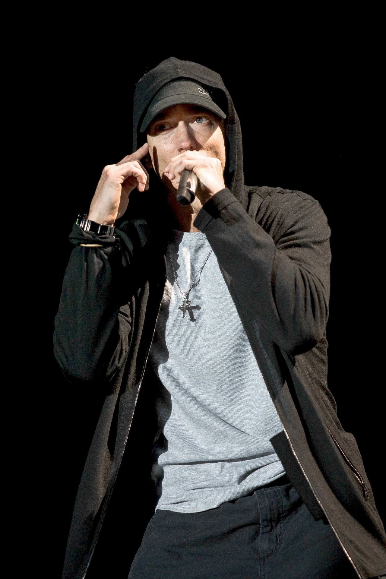 Eminem performing live with a microphone, wearing a black cap and hooded jacket
