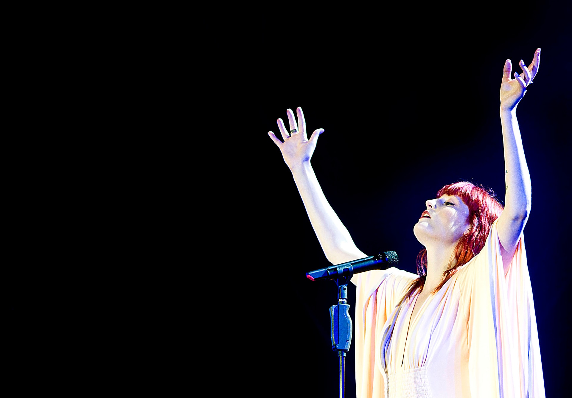 Florence Welch of Florence and the Machine performing on stage with both arms raised, lit in warm light against a black background