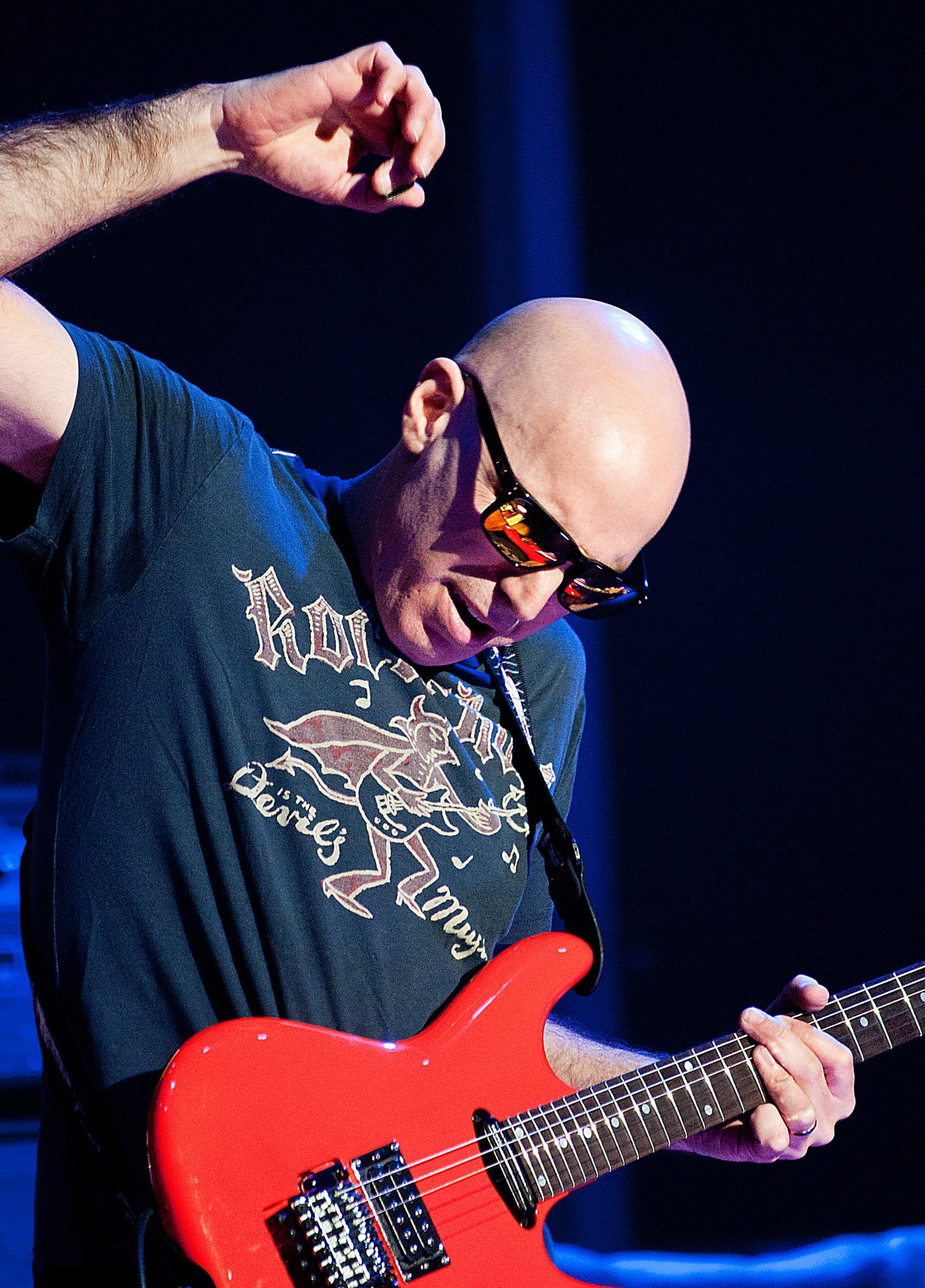 Joe Satriani playing a red electric guitar under blue stage lighting