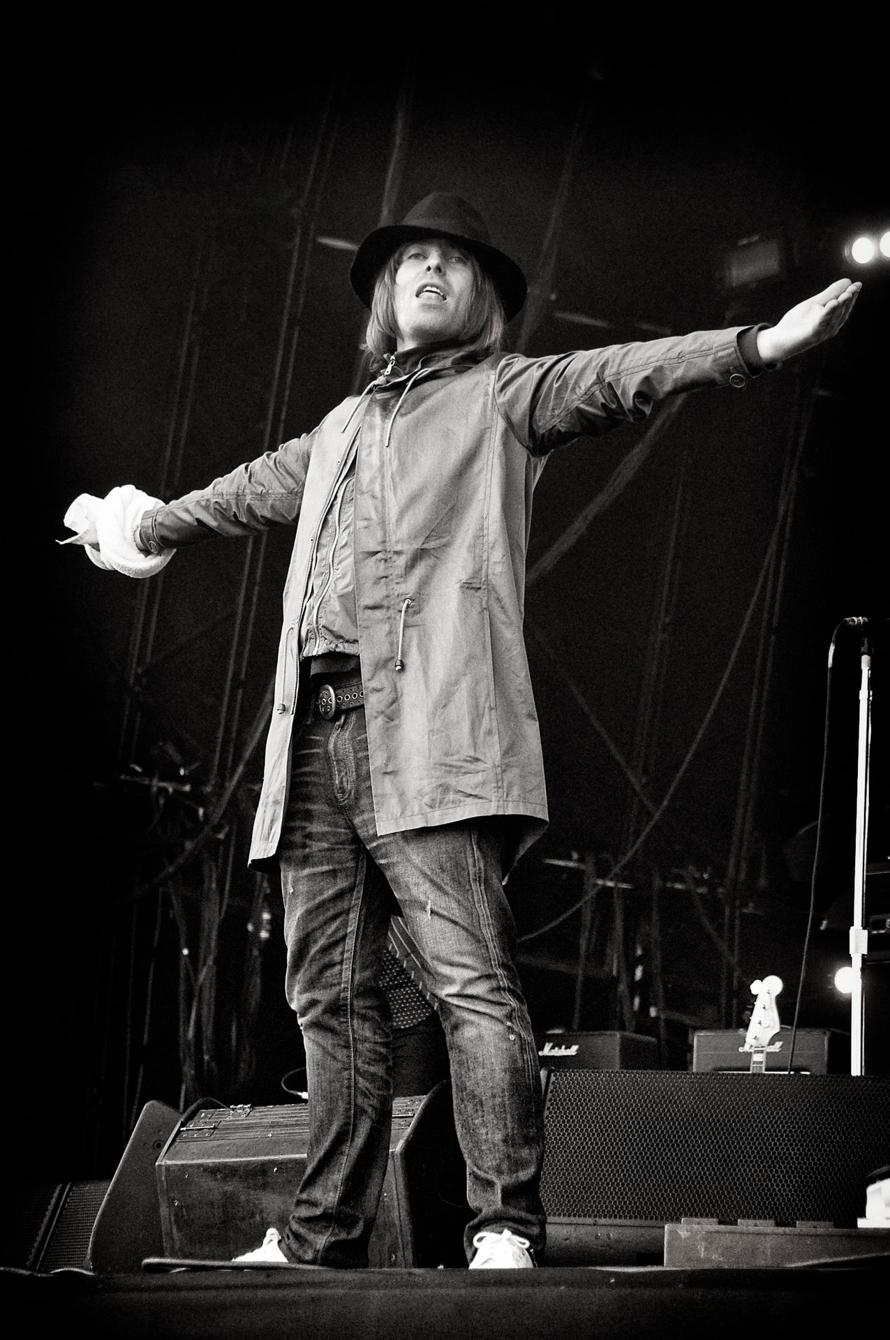 Liam Gallagher performing on stage in black and white, arms outstretched, wearing a long coat and wide-brimmed hat