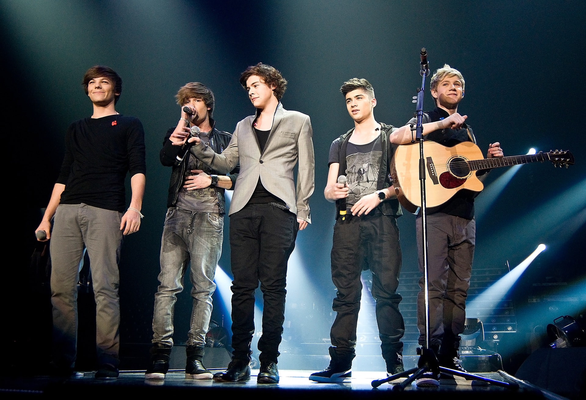 One Direction performing together on stage with microphones and acoustic guitar