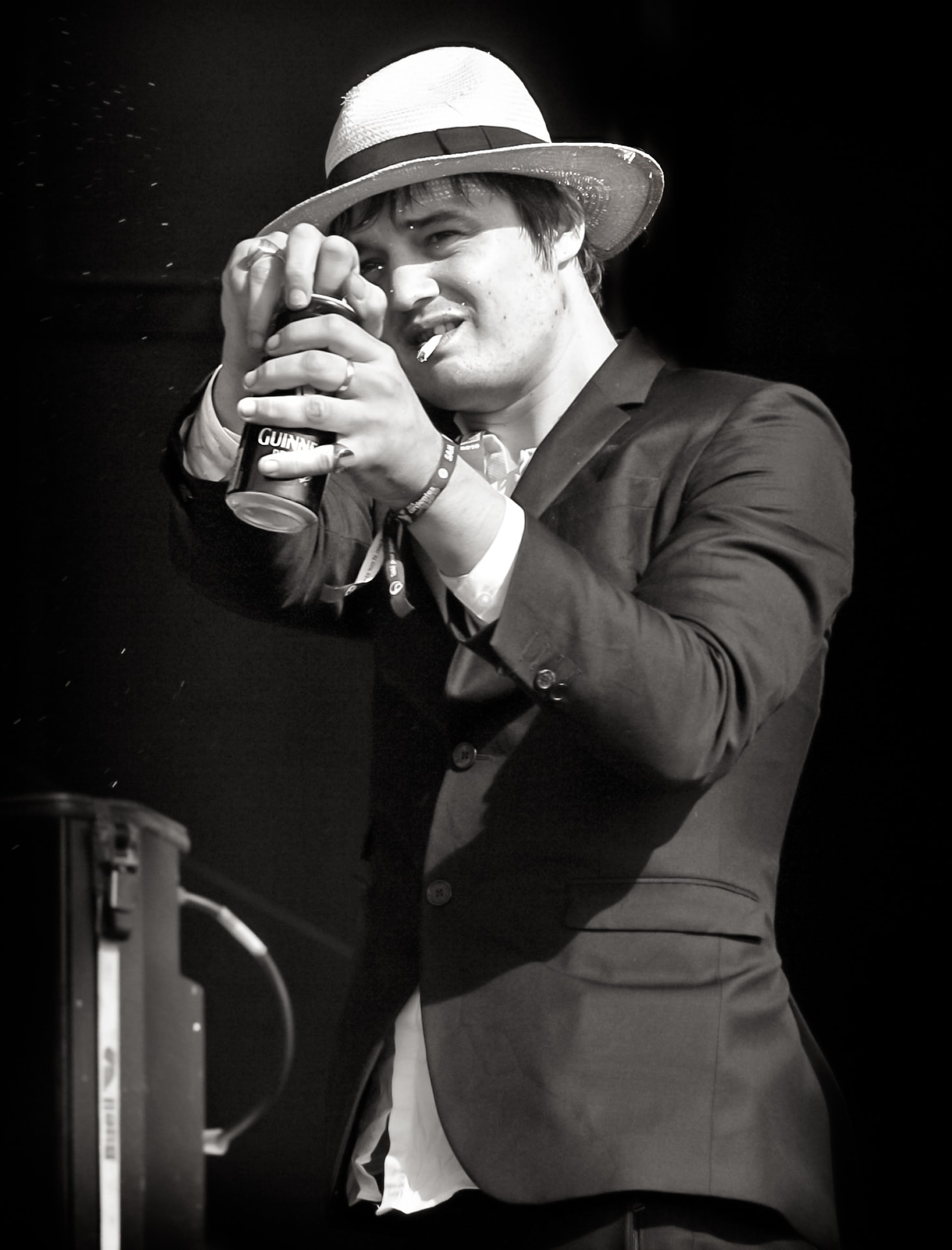 Pete Doherty on stage in a black-and-white portrait, wearing a hat and holding a drink