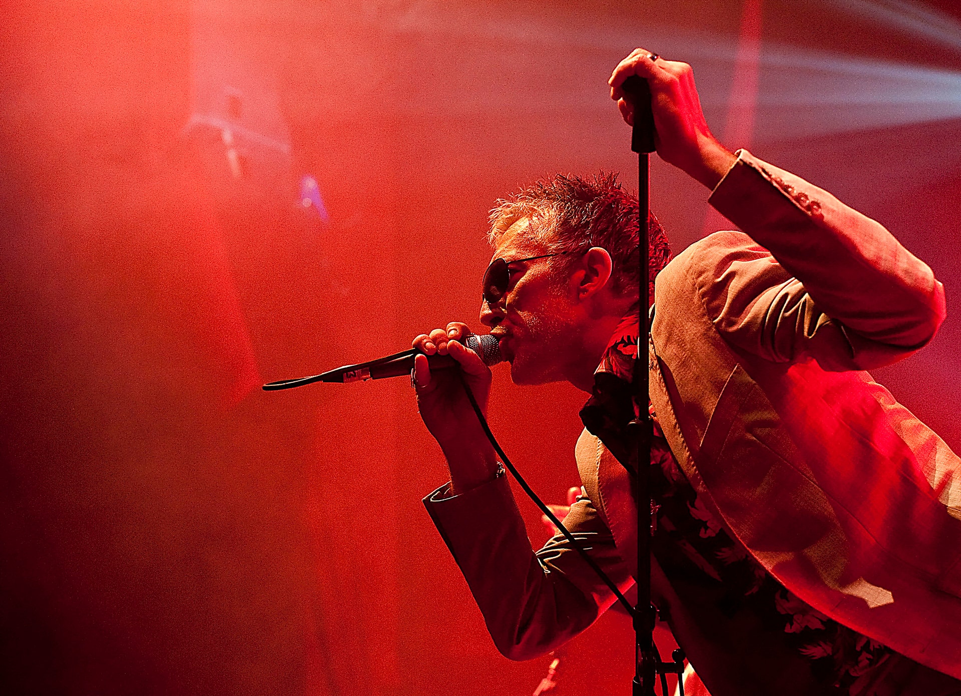 Rob Spragg of Alabama 3 singing into a microphone with fist raised under red stage lighting