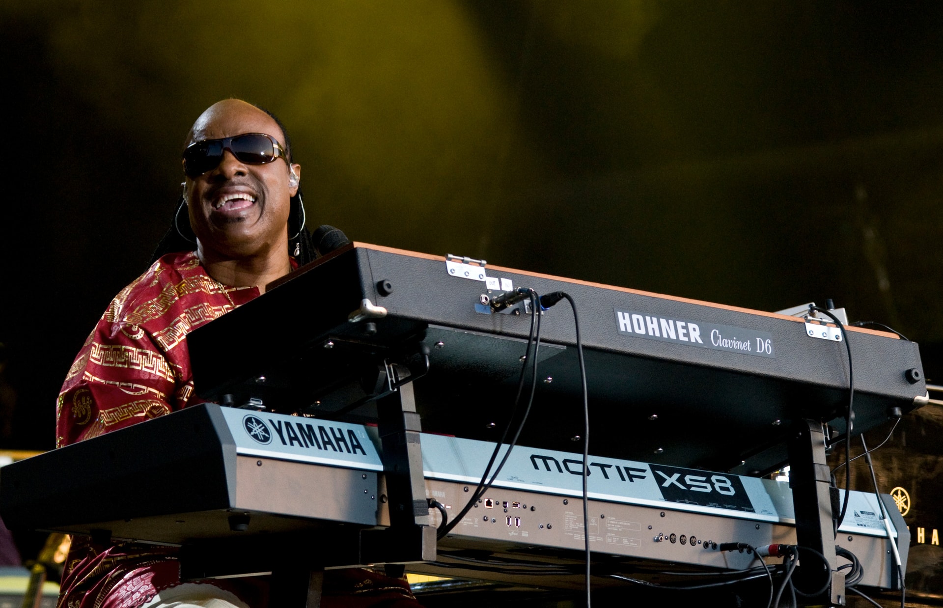 Stevie Wonder performing on stage behind a Yamaha keyboard, smiling, wearing a red and gold jacket